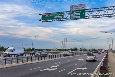 Ruch drogowy na wielopasmowej jezdni z tablicami kierunkowymi wskazującymi drogę S8/E67 do Białegostoku oraz zjazdy na ulice Modlińską i Jagiellońską.