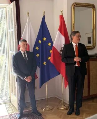 Dwaj mężczyźni w garniturach stoją w reprezentacyjnym pomieszczeniu przed flagami polską, unijną i austriacką.
