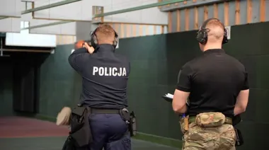 Policjant w strzelnicy oddaje strzał z pistoletu podczas treningu, obok stoi mężczyzna z notatnikiem.