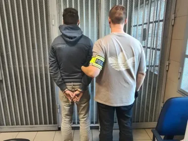 Funkcjonariusz policji trzyma zatrzymanego mężczyznę w kajdankach stojącego tyłem przed kratami w pomieszczeniu służbowym.