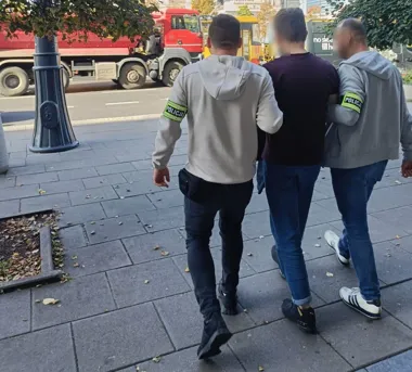 Funkcjonariusze policji w bluzach z opaskami "POLICJA" prowadzą zatrzymanego mężczyznę chodnikiem ulicy w ciągu dnia.