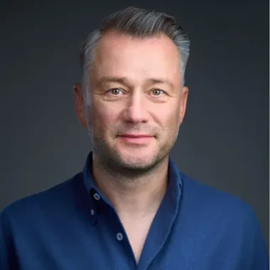 Portret mężczyzny w średnim wieku z siwiejącymi włosami, ubranego w granatową koszulę polo, na ciemnym tle.
