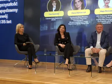 Trzy osoby siedzą na krzesłach na scenie podczas panelu dyskusyjnego, w tle wyświetlone są zdjęcia i nazwiska uczestników wydarzenia.