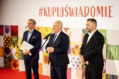Trzej mężczyźni w garniturach stoją na scenie podczas wydarzenia promocyjnego z hasłem #KUPUŚWIADOMIE na tle z kolorowymi grafikami żywności.