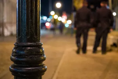 Fragment ozdobnego słupa na chodniku nocnej ulicy, w tle rozmyci przechodnie i światła latarni.