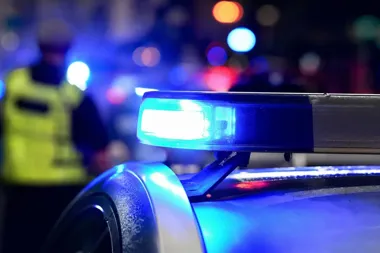 Nocna scena z policyjnym światłem błyskowym na dachu radiowozu, w tle rozmyta sylwetka funkcjonariusza w kamizelce odblaskowej.
