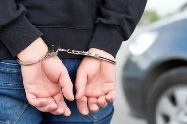 Zatrzymana osoba w czarnej bluzie i dżinsach stoi przy samochodzie z rękami skutymi kajdankami za plecami.