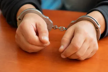 Skute kajdankami dłonie osoby w ciemnej bluzie leżą na drewnianym stole.