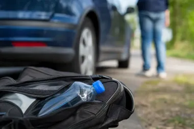 Czarny plecak z plastikową butelką leży na ziemi przy drodze, w tle stoi niebieskie auto i rozmyta postać człowieka.