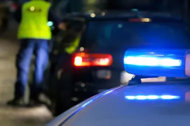 Policyjny radiowóz z włączonym niebieskim światłem podczas nocnej interwencji drogowej, w tle funkcjonariusz w kamizelce odblaskowej przy czarnym aucie.