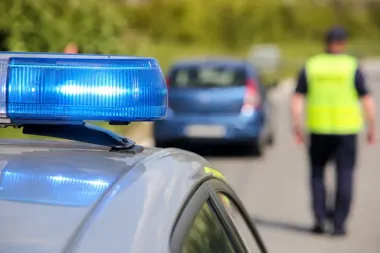 Policyjny radiowóz z niebieskim światłem na drodze, w tle funkcjonariusz w kamizelce odblaskowej i zatrzymany samochód osobowy.