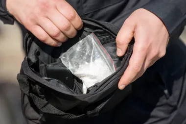 Mężczyzna w ciemnej kurtce otwiera czarną torbę, w której leży przezroczysty woreczek z białą substancją krysztaliczną.