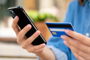 Osoba w niebieskiej koszuli trzyma smartfon i niebieską kartę płatniczą, wykonując operację online na zewnątrz.