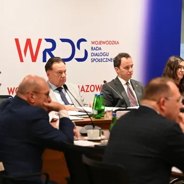 Uczestnicy posiedzenia Wojewódzkiej Rady Dialogu Społecznego w Mazowieckiem siedzą przy stole konferencyjnym z mikrofonami i dokumentami.