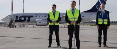 Trzej mężczyźni w kamizelkach odblaskowych stoją na płycie lotniska przed samolotem PLL LOT, jeden z nich przemawia do mikrofonu.