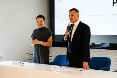 Mężczyzna w garniturze przemawia przez mikrofon obok stojącej kobiety podczas wydarzenia przy stole z tabliczkami nazwisk.