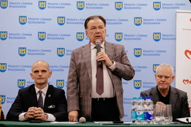 Mężczyzna w kraciastym garniturze przemawia do mikrofonu podczas konferencji w sali z logotypami Miasto i Gmina Bodzanów, przy stole siedzą dwaj inni mężczyźni.