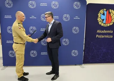 W pomieszczeniu przed ścianką Akademii Pożarniczej funkcjonariusz PSP w mundurze uroczyście wręcza dokument mężczyźnie w garniturze, który przyjmuje go uściskie