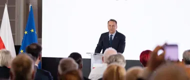 Mężczyzna w garniturze przemawia zza pulpitu do publiczności, obok flag Polski i Unii Europejskiej.