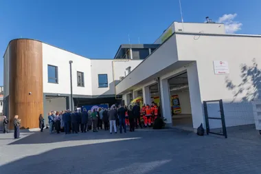 Grupa ludzi zgromadzona przed nowoczesnym budyniem służb ratunkowych, w którego garażach stoją ambulanse, podczas oficjalnego wydarzenia w słoneczny dzień.
