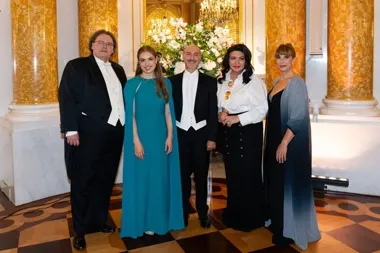 Pięć osób w strojach wieczorowych i smokingach pozuje do zdjęcia w reprezentacyjnym wnętrzu z kolumnami i żyrandolami.