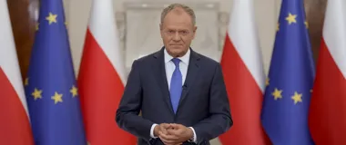 Mężczyzna w garniturze stoi przed flagami Polski i Unii Europejskiej w reprezentacyjnym wnętrzu.