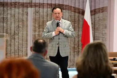 Mężczyzna w jasnej marynarce w kratę przemawia do siedzącej publiczności, trzymając mikrofon, w tle zasłona i polska flaga.