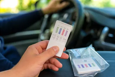 Kierowca w samochodzie trzyma w dłoni test do wykrywania narkotyków, a na desce rozdzielczej leży kolejny test w opakowaniu.