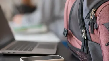 Plecak, laptop i smartfon leżące na stole w pomieszczeniu.