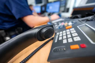 Słuchawka i panel telefonu leżą na biurku w centrum dyspozytorskim, w tle rozmyty pracownik przy komputerach.