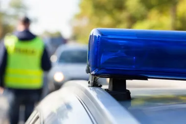 Policyjny kogut świetlny na dachu radiowozu, w tle rozmazany funkcjonariusz w kamizelce odblaskowej i samochód na drodze.
