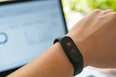 Nadgarstek osoby z czarną opaską fitness wyświetlającą czerwone ikony serca i EKG, w tle rozmyty ekran laptopa.