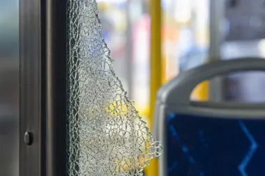 Zbite, pokruszone szkło w drzwiach lub oknie pojazdu komunikacji miejskiej, w tle widać siedzenie i fragmenty wnętrza autobusu lub tramwaju.
