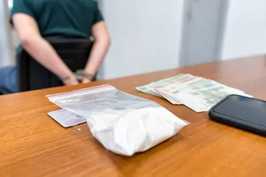 Zatrzymana osoba w kajdankach siedzi przy stole, na którym leży woreczek z białym proszkiem, banknoty, telefon i dokument.