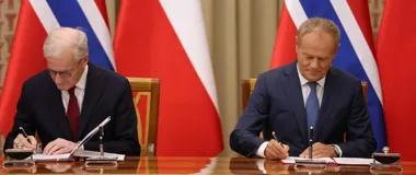 Dwaj mężczyźni w garniturach podpisują dokumenty przy drewnianym stole, za nimi flagi Polski i Norwegii.