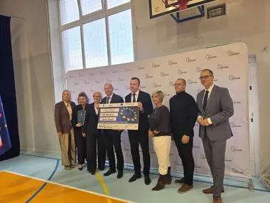 Grupa osób w hali sportowej Gminy Załuski pozuje z dużym czekiem na kwotę 543 660,00 zł z Funduszy Europejskich dla Mazowsza 2021-2027.