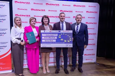 Grupa osób pozuje na scenie z dużym czekiem dotacyjnym na rewitalizację Pałacu Biskupów Płockich w Broku podczas uroczystości w Mazowszu.