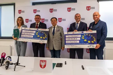 Pięcioro osób w sali konferencyjnej w Płońsku trzyma duże czeki z funduszy europejskich na tle ścianki z herbem i napisem Miasto Płońsk.