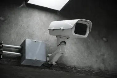 Biała kamera monitoringu zamontowana na betonowej ścianie obok skrzynki elektrycznej z rurami.