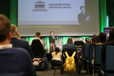 Kobieta przemawia z mikrofonem na scenie podczas kongresu organizowanego przez Rzecznika Praw Obywatelskich, wśród publiczności osoby na wózkach inwalidzkich.