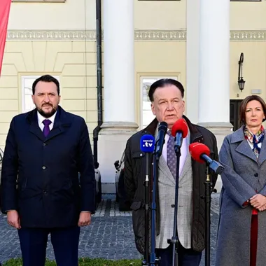 Trzy osoby stoją przed kolumnami budynku, mężczyzna w środku przemawia do mikrofonów dziennikarskich.