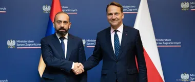 Dwaj mężczyźni w garniturach witają się uściskiem dłoni podczas spotkania w Ministerstwie Spraw Zagranicznych, w tle flagi Armenii i Polski.