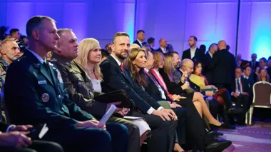 Uczestnicy oficjalnej uroczystości lub konferencji siedzą w pierwszych rzędach sali oświetlonej fioletowym światłem.