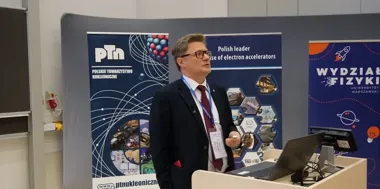 Mężczyzna w garniturze wygłasza prezentację przy pulpicie z laptopem, w tle banery Polskiego Towarzystwa Nukleonicznego i Wydziału Fizyki UW.