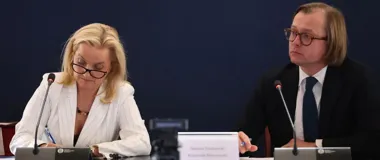 Dwie osoby siedzą przy stole podczas formalnego spotkania lub konferencji; kobieta w białym prążkowanym garniturze robi notatki, a mężczyzna w czarnym garniturz
