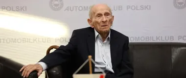 Starszy mężczyzna w garniturze siedzi na czarnej kanapie podczas wydarzenia Automobilklubu Polskiego.