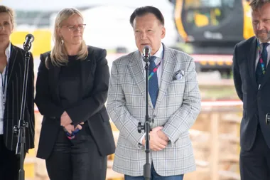 Cztery osoby stoją przy mikrofonach na zewnątrz, prawdopodobnie podczas konferencji prasowej lub wydarzenia oficjalnego na placu budowy.