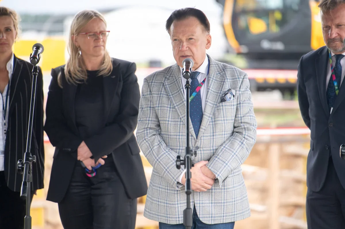 Cztery osoby stoją przy mikrofonach na zewnątrz, prawdopodobnie podczas konferencji prasowej lub wydarzenia oficjalnego na placu budowy.
